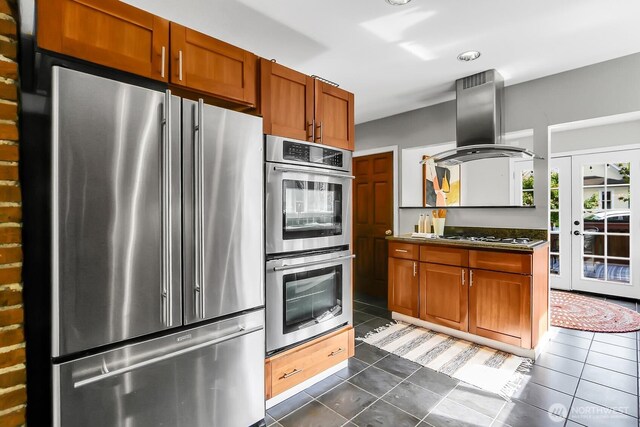 kitchen with appliances with stainless steel finishes, island exhaust hood, dark tile patterned flooring, and brown cabinetry