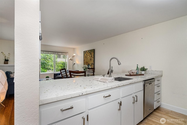 kitchen with stainless steel dishwasher, a sink, light wood finished floors, white cabinetry, and light stone countertops