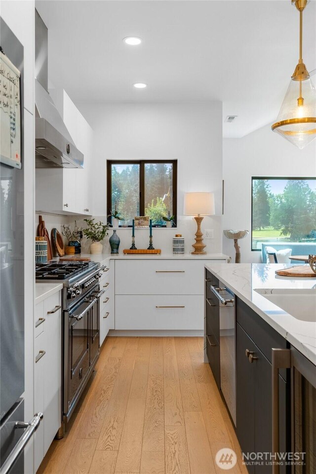 kitchen with double oven range, wall chimney exhaust hood, light wood finished floors, white cabinetry, and a sink