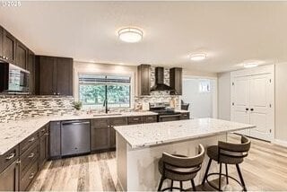 kitchen featuring wall chimney range hood, stainless steel appliances, light wood-style flooring, a kitchen island, and a kitchen breakfast bar