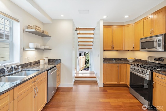 kitchen with appliances with stainless steel finishes, a sink, light wood-type flooring, open shelves, and dark stone counters