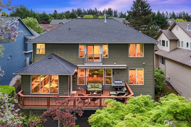 back of house at dusk featuring a shingled roof, a balcony, and a deck