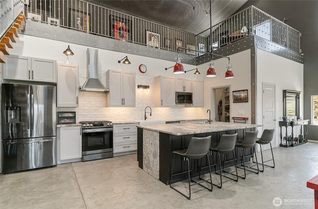 kitchen with appliances with stainless steel finishes, wall chimney range hood, a towering ceiling, concrete floors, and decorative backsplash