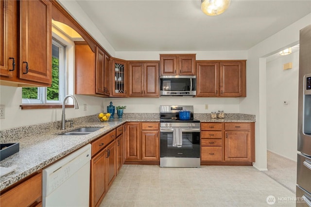 kitchen with stainless steel appliances, a sink, light stone counters, brown cabinets, and glass insert cabinets