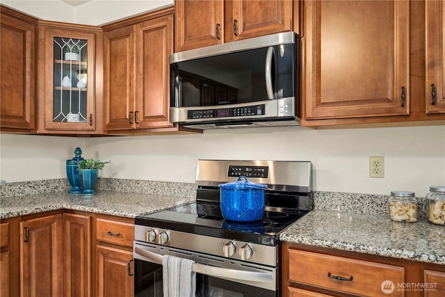 kitchen featuring appliances with stainless steel finishes, brown cabinetry, glass insert cabinets, and light stone counters