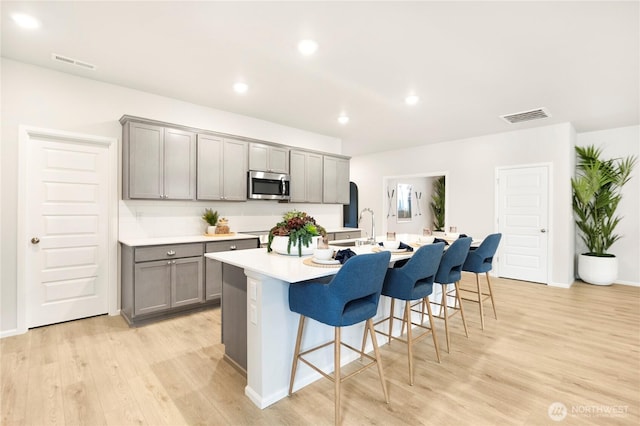 kitchen featuring gray cabinetry, stainless steel microwave, light wood-style flooring, a center island with sink, and light countertops