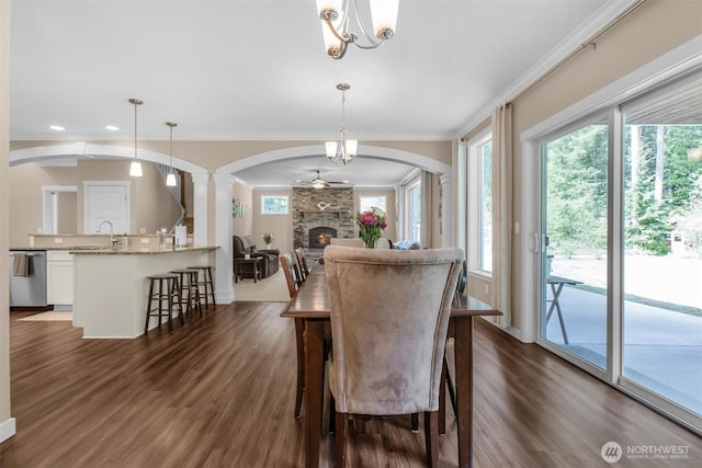 dining area featuring arched walkways, dark wood-type flooring, a chandelier, ornamental molding, and a stone fireplace