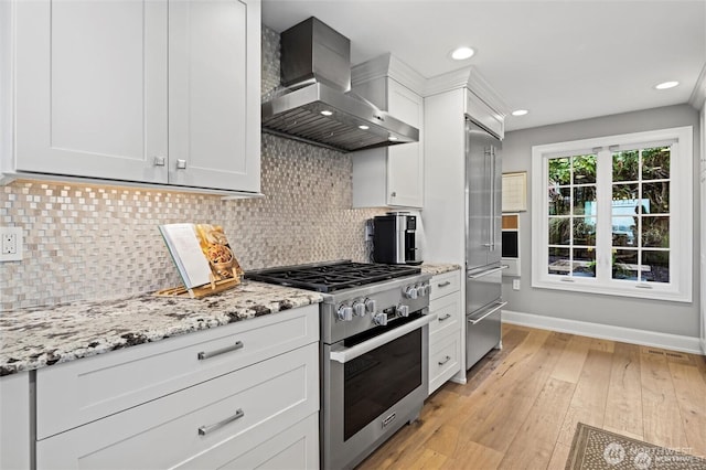 kitchen featuring wall chimney exhaust hood, high end appliances, baseboards, white cabinetry, and light stone counters
