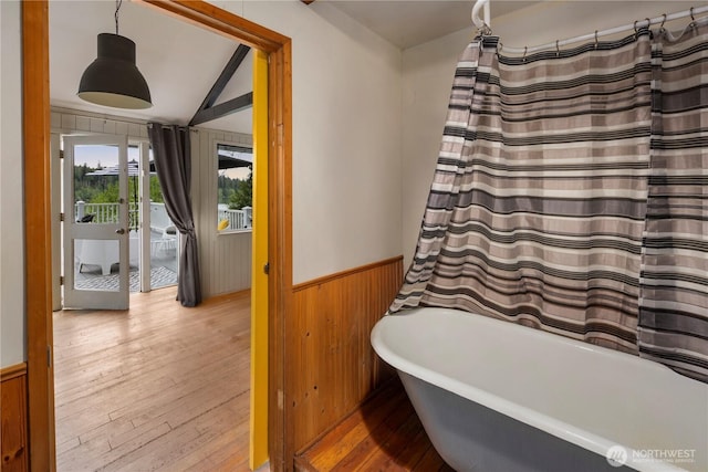 bathroom with a wainscoted wall, wood walls, wood-type flooring, a bathtub, and vaulted ceiling
