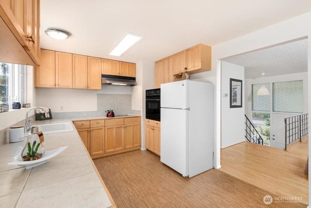 kitchen featuring black appliances, under cabinet range hood, light brown cabinets, a sink, and tasteful backsplash