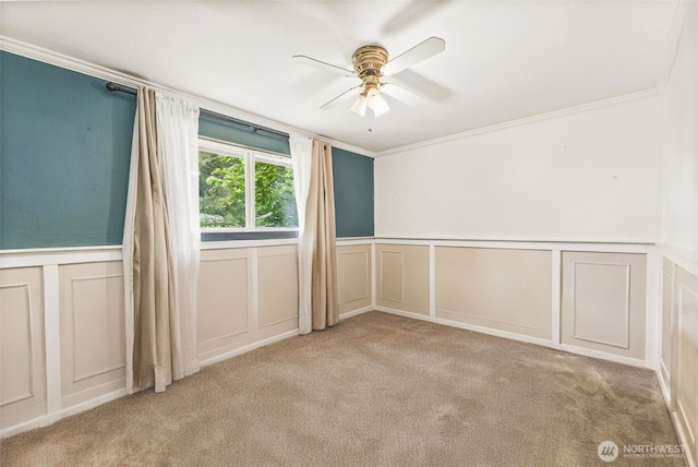 unfurnished room featuring a decorative wall, a ceiling fan, carpet flooring, crown molding, and wainscoting