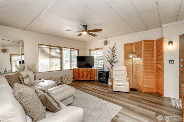 living area with ceiling fan, wood finished floors, a wood stove, and ornamental molding