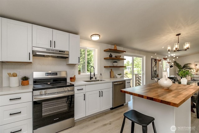 kitchen featuring under cabinet range hood, appliances with stainless steel finishes, butcher block countertops, a sink, and a chandelier
