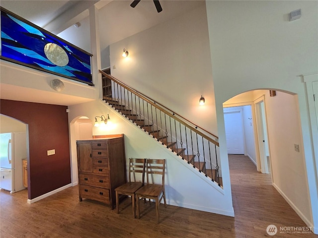 stairs featuring a towering ceiling, ceiling fan, arched walkways, wood finished floors, and baseboards