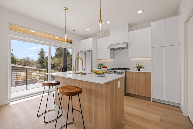 6730 21st Ave SW, Seattle, WA 98106 - kitchen with wall chimney range hood, appliances with stainless steel finishes, modern cabinets, a center island with sink, and light wood-style flooring