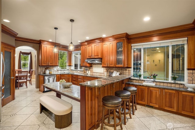 24702 64th Ave E, Graham, WA 98338 - kitchen featuring stainless steel dishwasher, brown cabinetry, under cabinet range hood, a kitchen breakfast bar, and crown molding