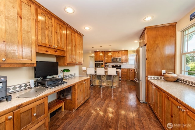 516 E Willow St, Sequim, WA 98382 - kitchen with stainless steel appliances, brown cabinetry, decorative light fixtures, dark wood-style flooring, and a desk