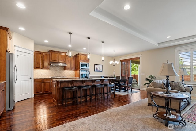 4837 Castleton Rd SW, Port Orchard, WA 98367 - kitchen with appliances with stainless steel finishes, under cabinet range hood, dark wood-style flooring, a breakfast bar, and brown cabinets