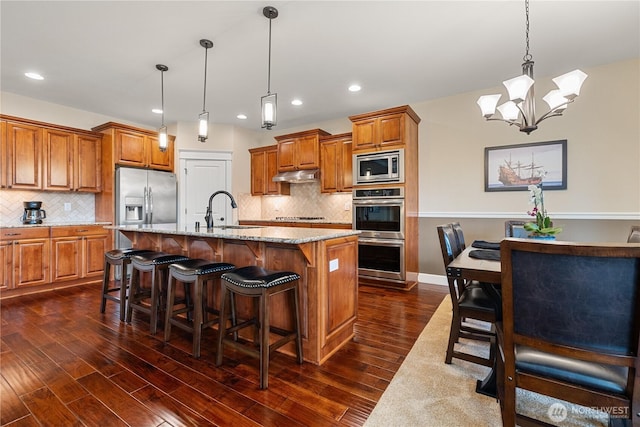 4837 Castleton Rd SW, Port Orchard, WA 98367 - kitchen with appliances with stainless steel finishes, under cabinet range hood, brown cabinetry, light stone countertops, and a chandelier