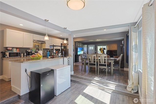 373 Adams Ave, Morton, WA 98356 - kitchen with under cabinet range hood, white cabinetry, light wood-style flooring, freestanding refrigerator, and an island with sink