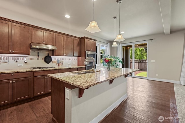 8910 Buttercup St SE, Tumwater, WA 98501 - kitchen with under cabinet range hood, stainless steel appliances, a center island with sink, dark wood-style flooring, and light stone countertops