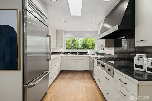 3015 44th Ave W, Seattle, WA 98199 - kitchen featuring stainless steel appliances, wall chimney exhaust hood, white cabinetry, light wood-type flooring, and a skylight