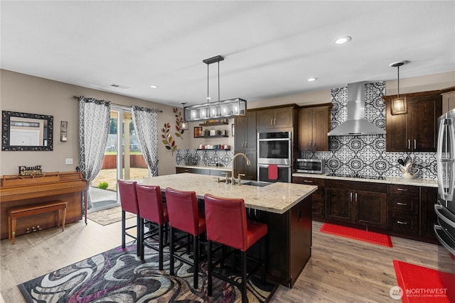 22817 80th Avenue Ct E, Graham, WA 98338 - kitchen with wall chimney range hood, appliances with stainless steel finishes, light wood-style flooring, tasteful backsplash, and a center island with sink