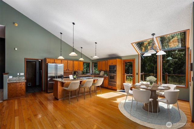 2515 87th Avenue Ct E, Edgewood, WA 98371 - kitchen with appliances with stainless steel finishes, brown cabinetry, high vaulted ceiling, light wood finished floors, and a center island with sink