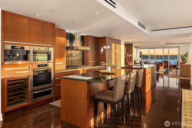 99 Union St Unit 1601, Seattle, WA 98101 - kitchen with wall chimney range hood, a warming drawer, dark wood-style floors, brown cabinets, and appliances with stainless steel finishes