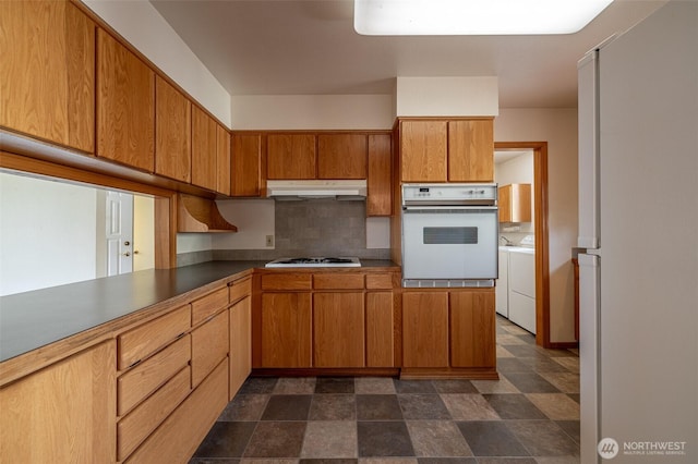 1312 Welch Ave, Wenatchee, WA 98801 - kitchen featuring white appliances, under cabinet range hood, tasteful backsplash, washing machine and dryer, and brown cabinetry