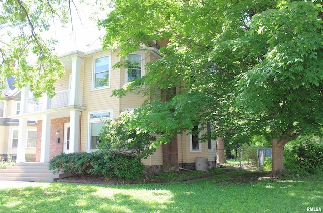 view of property hidden behind natural elements with a front lawn, a balcony, and central air condition unit