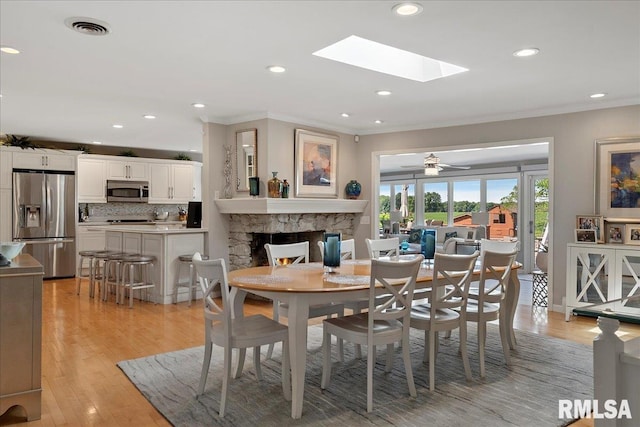 dining room with light wood-type flooring, a skylight, ceiling fan, crown molding, and a stone fireplace