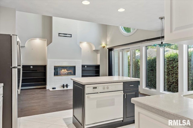 kitchen with white oven, light wood-type flooring, a tiled fireplace, stainless steel refrigerator, and white cabinets