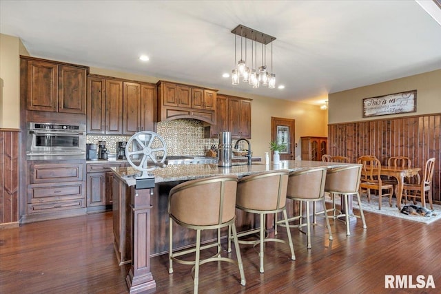 kitchen with decorative light fixtures, a kitchen island with sink, dark wood-type flooring, stainless steel oven, and decorative backsplash