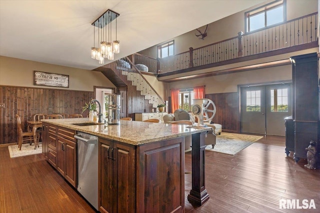 kitchen with an island with sink, dark hardwood / wood-style flooring, pendant lighting, stainless steel dishwasher, and wooden walls