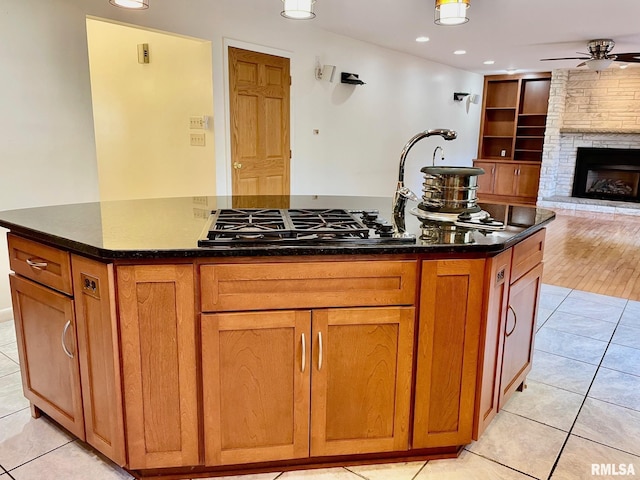 kitchen featuring a stone fireplace, built in features, a kitchen island with sink, light tile patterned floors, and ceiling fan