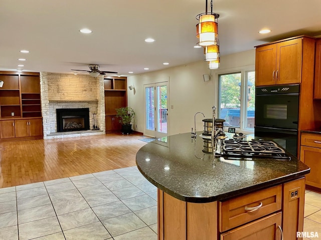 kitchen with pendant lighting, double oven, an island with sink, dark stone counters, and gas cooktop