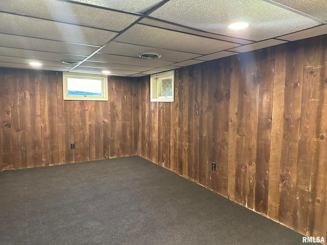 basement with a drop ceiling, wooden walls, and dark colored carpet