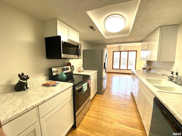 kitchen with a raised ceiling, white cabinetry, sink, stainless steel appliances, and light wood-type flooring