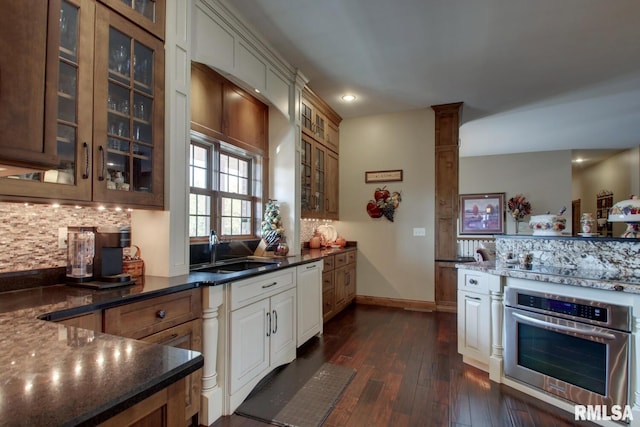 kitchen with stainless steel oven, dark wood-type flooring, dark stone countertops, sink, and tasteful backsplash