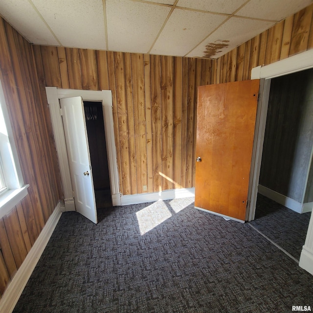 unfurnished room featuring wood walls, a drop ceiling, and dark carpet