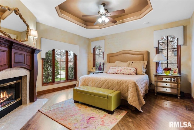 bedroom featuring a tray ceiling, a tiled fireplace, ceiling fan, and light hardwood / wood-style flooring