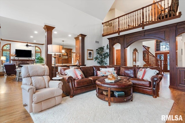 living room featuring high vaulted ceiling, a brick fireplace, ceiling fan, light wood-type flooring, and decorative columns