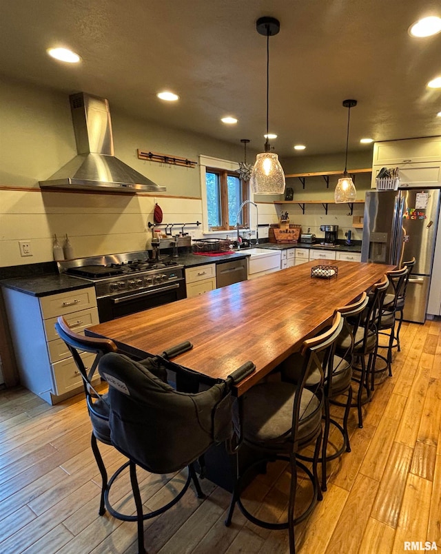 bar featuring light hardwood / wood-style flooring, wall chimney exhaust hood, appliances with stainless steel finishes, butcher block countertops, and decorative light fixtures