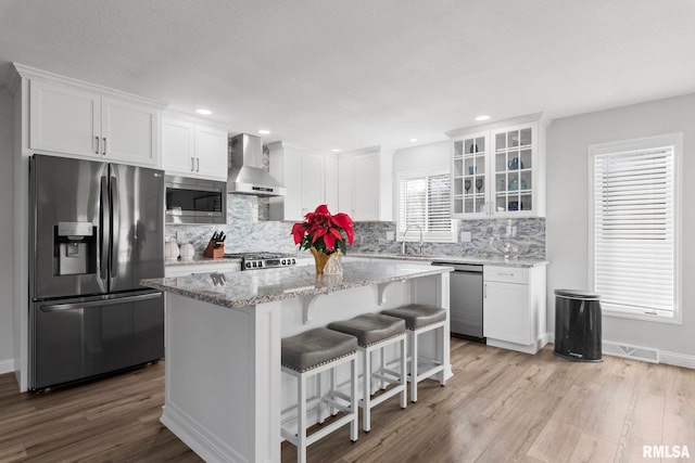 kitchen with a center island, stainless steel appliances, wall chimney range hood, white cabinets, and light wood-type flooring