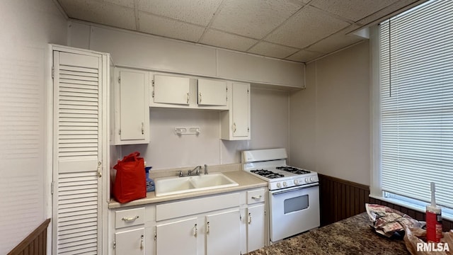 kitchen featuring a drop ceiling, white range with gas cooktop, wooden walls, sink, and white cabinets