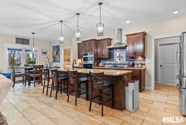 kitchen featuring appliances with stainless steel finishes, pendant lighting, an island with sink, decorative backsplash, and wall chimney exhaust hood