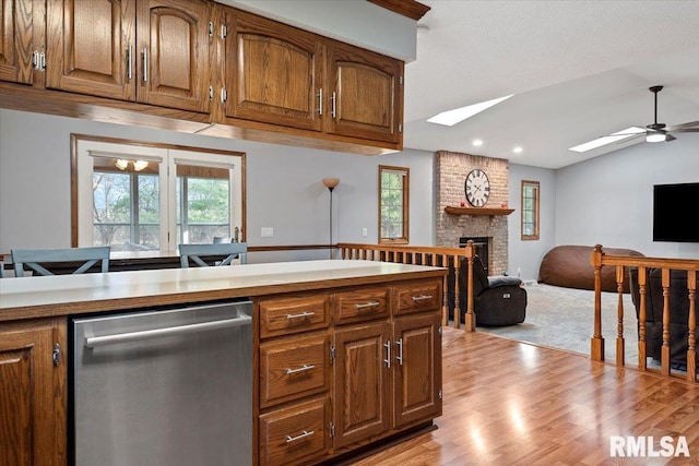 kitchen featuring ceiling fan, lofted ceiling with skylight, a fireplace, light hardwood / wood-style floors, and stainless steel dishwasher