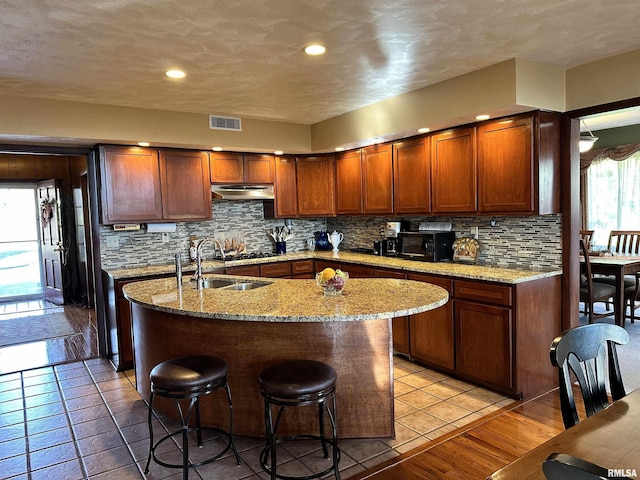 kitchen with tasteful backsplash, sink, a kitchen breakfast bar, a kitchen island with sink, and light stone countertops