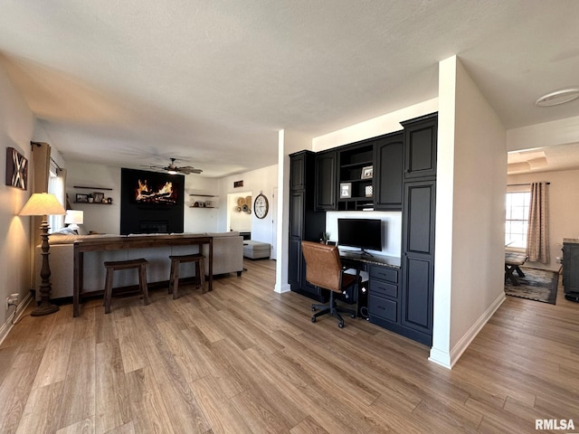 home office with light wood finished floors, a large fireplace, baseboards, a ceiling fan, and built in study area